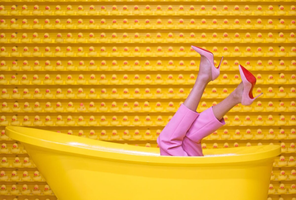 about-us-01 Legs in pink pants and red high heels pop out of a yellow bathtub against a creative rubber duck wall.